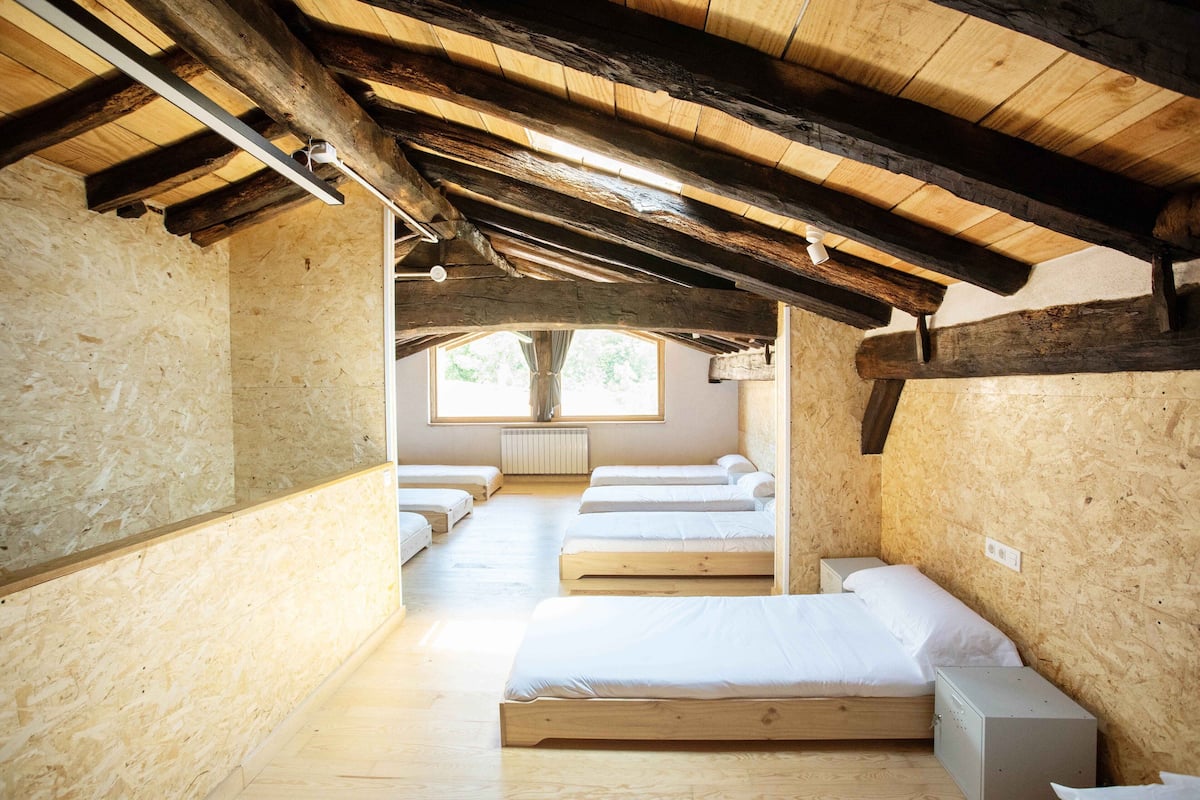 A wooden interior with bunk beds arranged in a row, a window with a view, and a radiator on the wall.