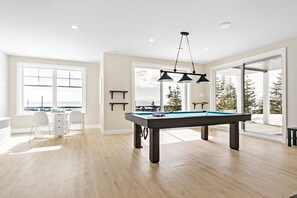 Una habitación amplia con mesa de billar, comedor con sillas y grandes ventanales que ofrecen vistas a árboles y un paisaje nevado.