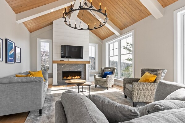 A spacious living room with a fireplace, large windows, and a high ceiling.