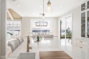 A spacious living room with a large window offering a scenic view, a dining area with a white table and chairs, and a modern kitchen with white cabinetry and a marble countertop.