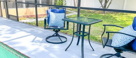 A poolside area with a glass table and two chairs, one with a patterned cushion.
