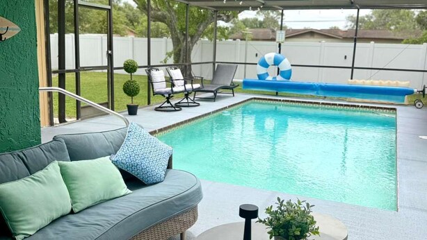 A covered outdoor area with a pool, a gray sofa with cushions, a round table with a plant, and lounge chairs. - Entire home with Screened- In Pool , Fully Fenced. (Spring Hill)