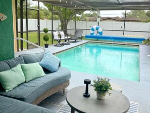A covered outdoor area with a pool, a gray sofa with cushions, a round table with a plant, and lounge chairs.