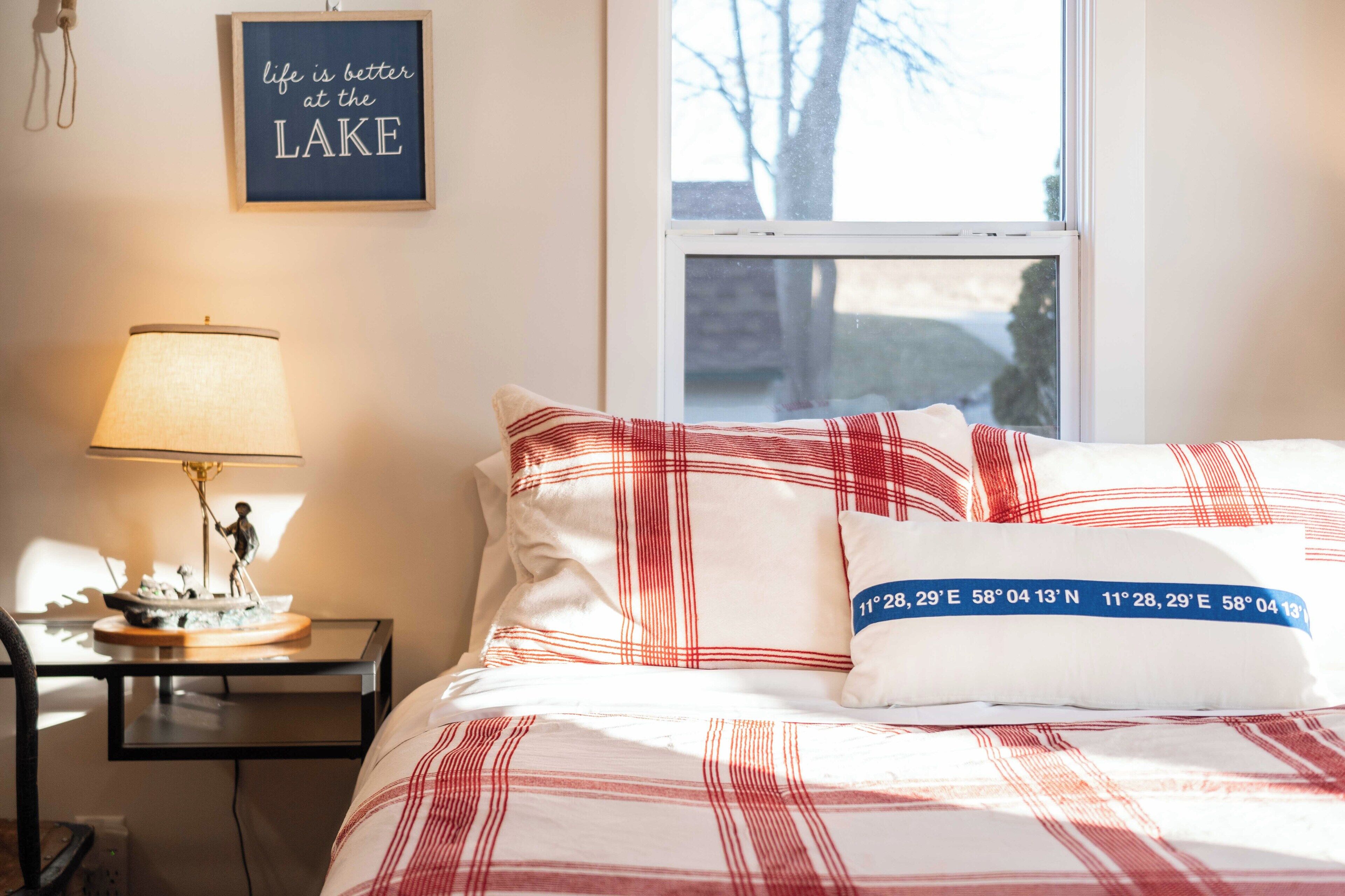 Un lit bien fait, avec une literie rouge et blanche, une table de chevet avec une lampe et une petite figurine de bateau, et un panneau mural sur lequel on peut lire : « La vie est plus belle au bord du LAC ».