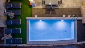 Una piscina exterior con sillones de descanso y una mesa, rodeada por un muro de piedra y una zona verde.