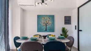 Un comedor moderno con una mesa dispuesta para cuatro personas, rodeada de sillas. Un cuadro de un árbol adorna la pared, y hay una planta en maceta a un lado.