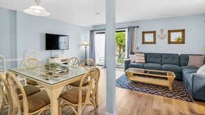 A modern living room with a grey sofa, a glass dining table, wicker chairs, and a flat-screen TV. - ⛱️Walk to Beach- Pool- Southbay#88 Townhome☀️ (Destin)