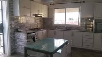 A modern kitchen with white cabinets, a glass-top table, and a window with blinds.