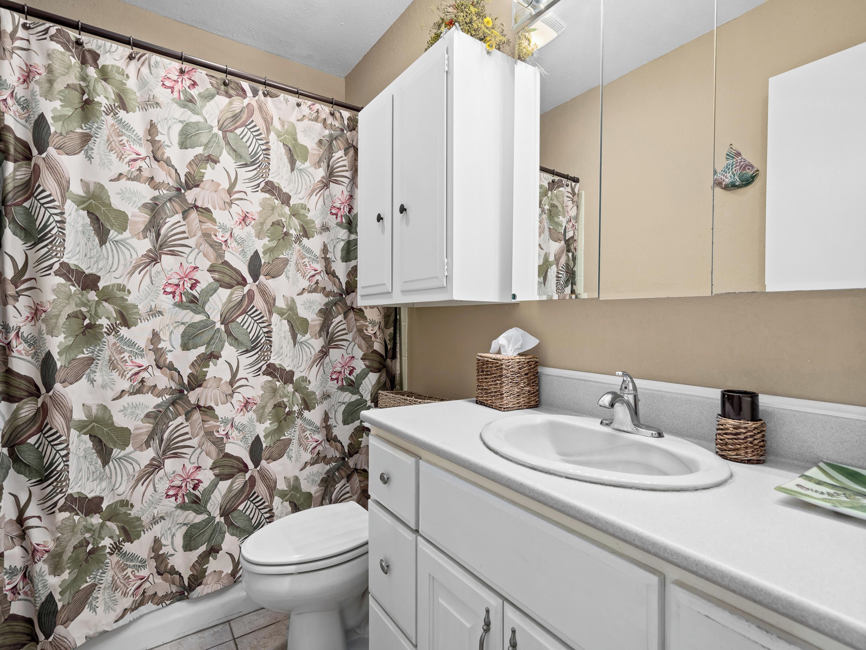 A bathroom with a toilet, a sink, and a mirror. There is a floral wallpaper on the wall, and a basket with a tissue on the sink counter.