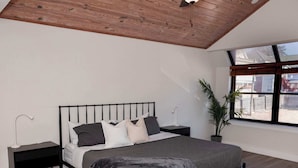 Een slaapkamer met een schuine plafond, een bed met hoofdbord, een nachtkastje, een vloerlamp, een potplant en een raam.