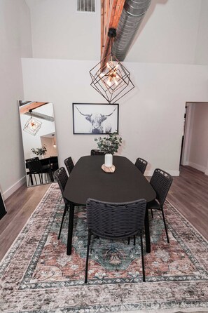 Un área de comedor con una mesa oscura, sillas negras, un tapete estampado, una lámpara colgante y un cuadro en la pared.