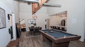 Een moderne woonkamer met een pooltafel, een eethoek, en een keuken die zichtbaar is door een open deur.