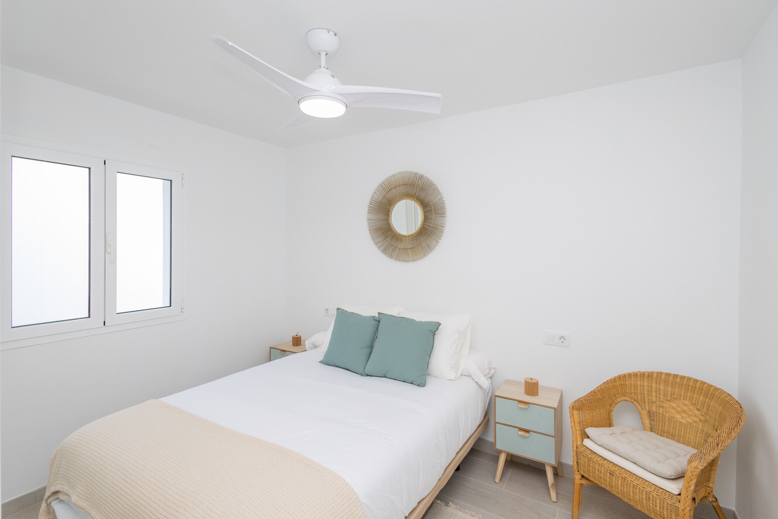 A bedroom with a bed, nightstands, a wicker chair, a ceiling fan, and a wall-mounted mirror.