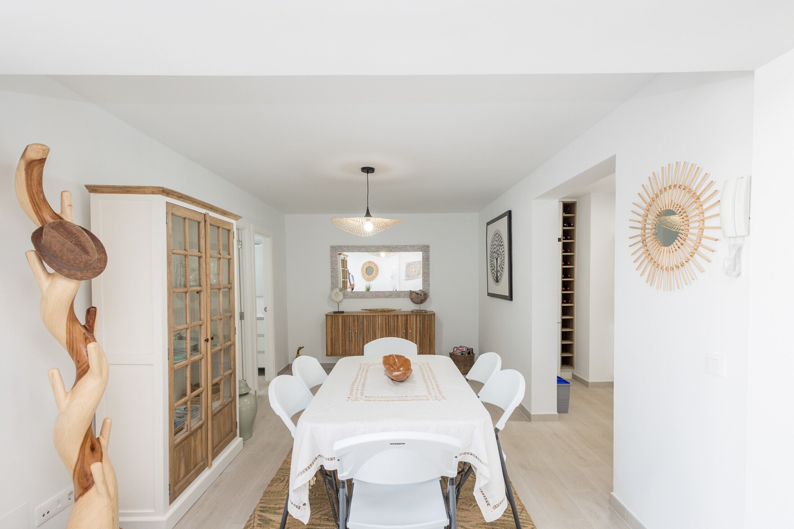 A dining area with a white table, chairs, and a central decorative bowl. There is a wooden cabinet with glass doors and a hanging light fixture above the table.