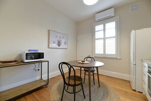 Et lille køkken med mikroovn, spisebord med stole, køleskab og et vindue med persienner.