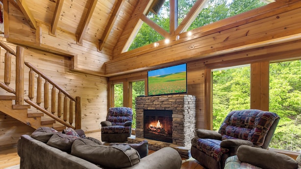 A log cabin interior with a stone fireplace, two armchairs, a sofa, and a staircase. - Mountain Cinema Lodge by Jackson Mountain Rentals (Sevierville)