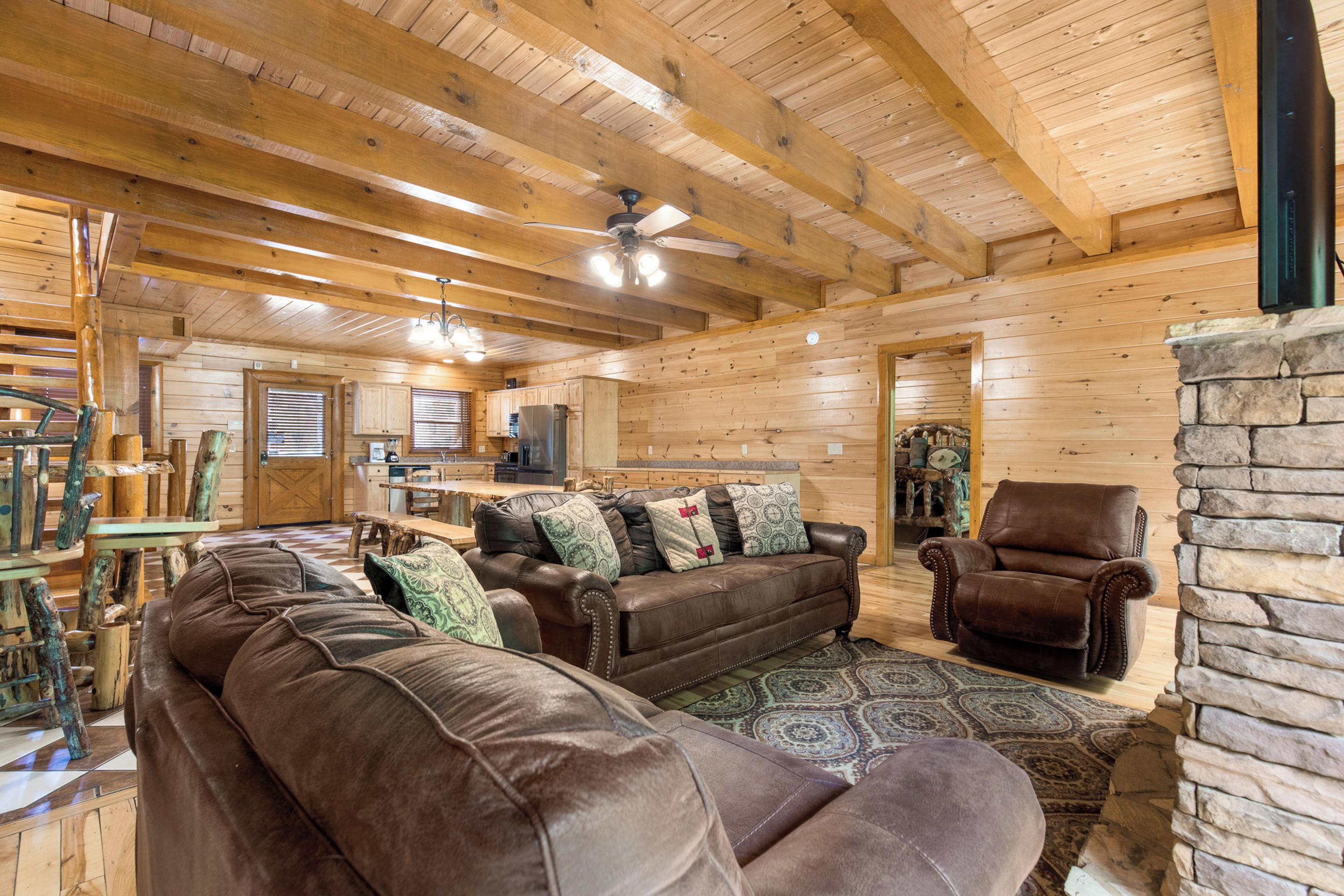 A cozy log cabin interior with a brown leather sofa, a stone fireplace, and a wooden ceiling with exposed beams.