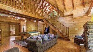 A spacious wooden cabin interior with a pool table, a sofa, a staircase, and a stone fireplace. - Mountain Cinema Lodge by Jackson Mountain Rentals (Sevierville)