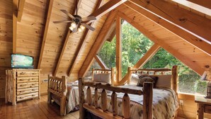 A wooden cabin bedroom with a loft bed, a television, and a large window overlooking greenery. - Mountain Cinema Lodge by Jackson Mountain Rentals (Sevierville)