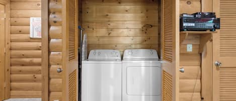 A laundry room with two white washing machines, wooden walls, and a shelf with electronic devices.