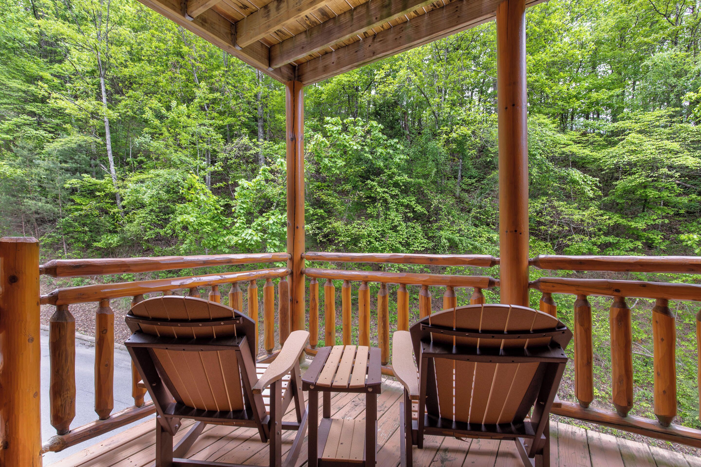 A wooden deck with two Adirondack chairs and a table, overlooking a forested area.