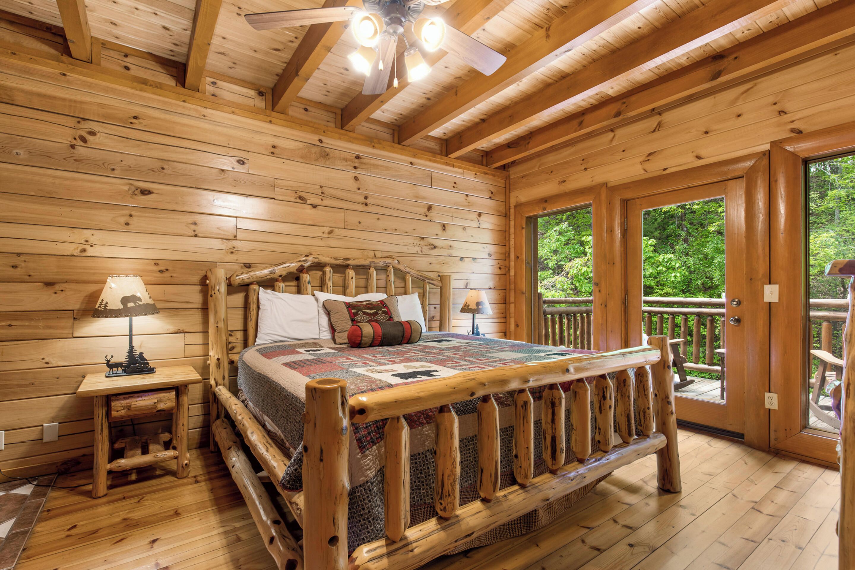 A wooden cabin bedroom with a bed, two bedside tables, a ceiling fan, and a balcony with a wooden railing.