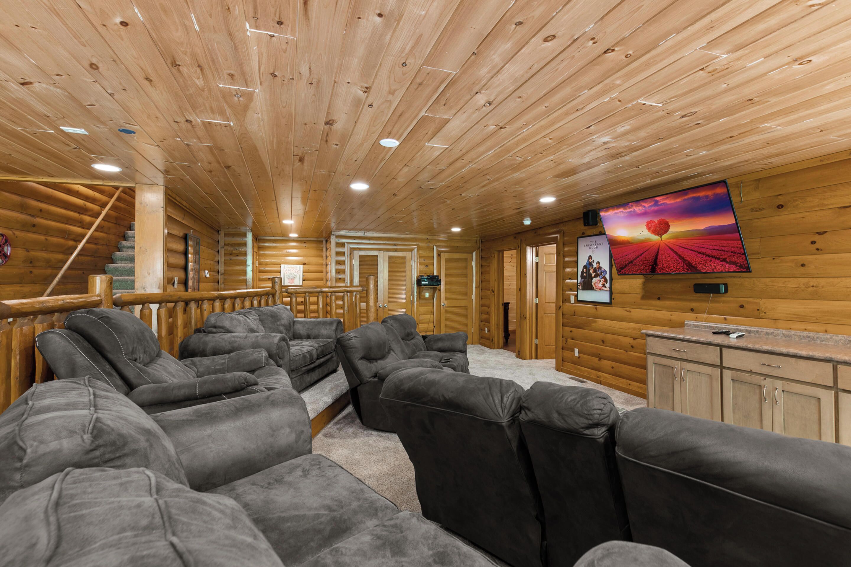 A log cabin interior with a large gray sectional sofa, wooden walls, and a mounted TV displaying a sunset scene.