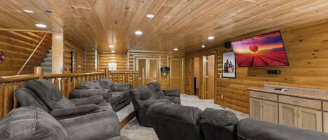 A log cabin interior with a large gray sectional sofa, wooden walls, and a mounted TV displaying a sunset scene.