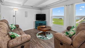 A cozy living room with a sofa set, a television, and a view of a grassy area through large windows. - Little Cove Resort by Miss Kitty’s (Taft)