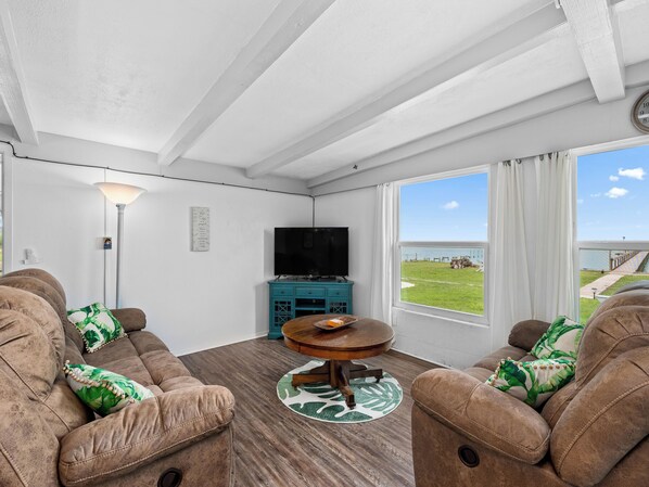 A cozy living room with a sofa set, a television, and a view of a grassy area through large windows. - Little Cove Resort by Miss Kitty’s (Taft)
