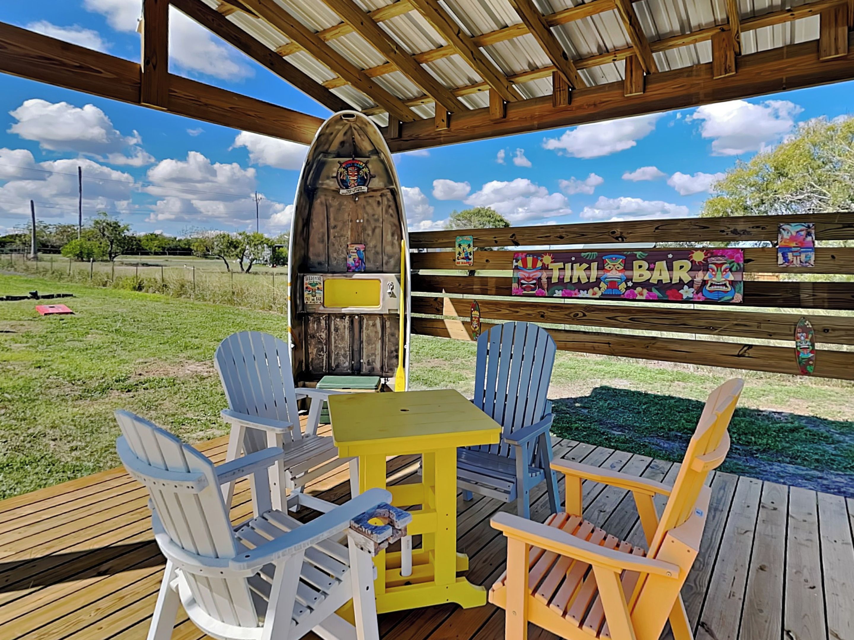 A covered outdoor seating area with wooden chairs and a table, a tiki bar sign, and a wooden structure in the background.