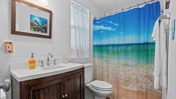 Une salle de bain avec un rideau de douche représentant une scène de plage, un lavabo avec un distributeur de savon et des toilettes.