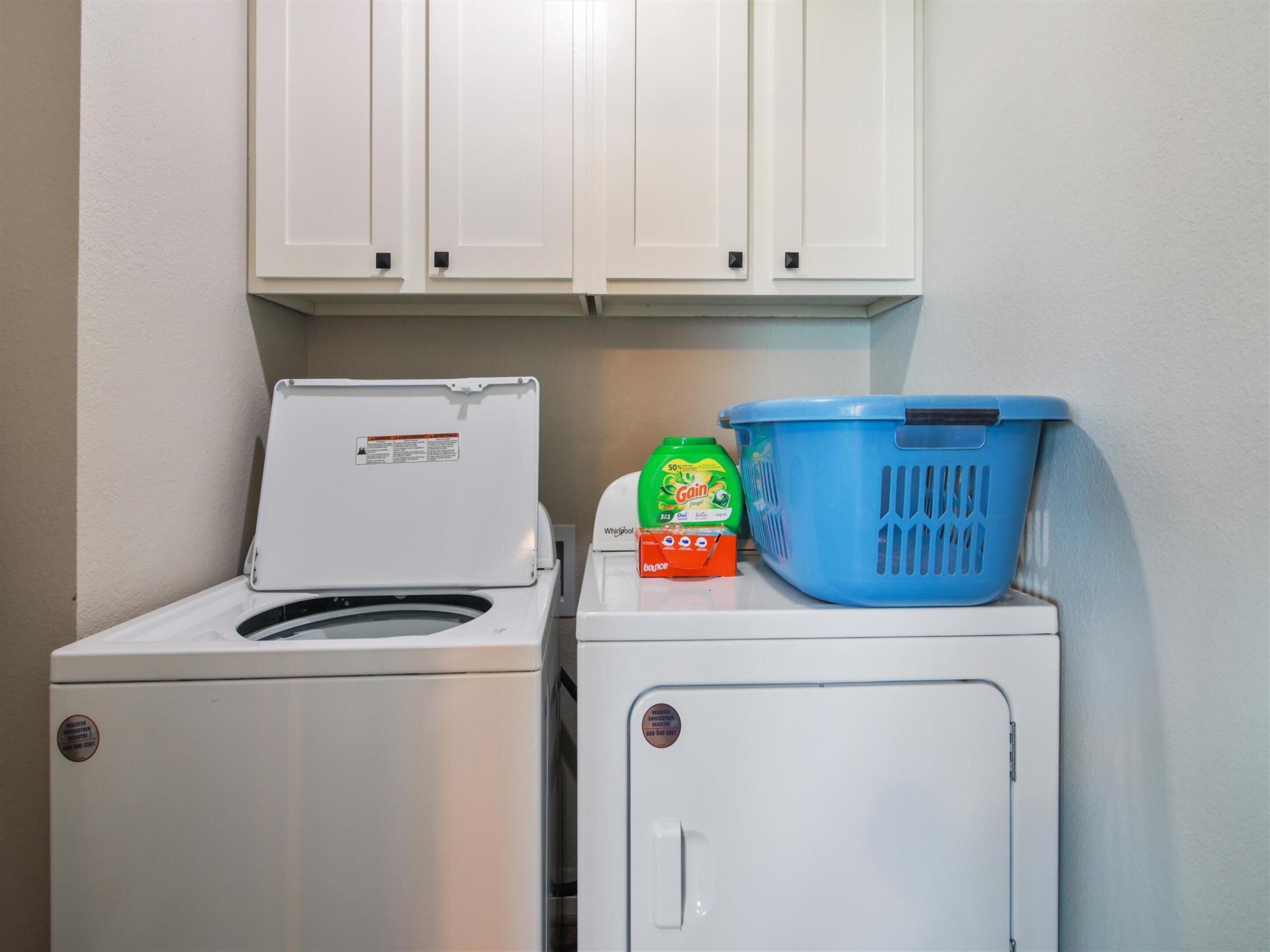 Un espace buanderie équipé d’un lave-linge et d’un sèche-linge à chargement frontal, d’un panier à linge bleu et d’une bouteille de lessive Gain.