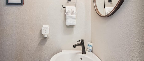 Un bagno con un lavandino bianco, uno specchio, un porta asciugamani e una presa di corrente.