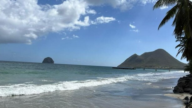 Una playa con cielos despejados, olas tranquilas y una montaña a lo lejos.