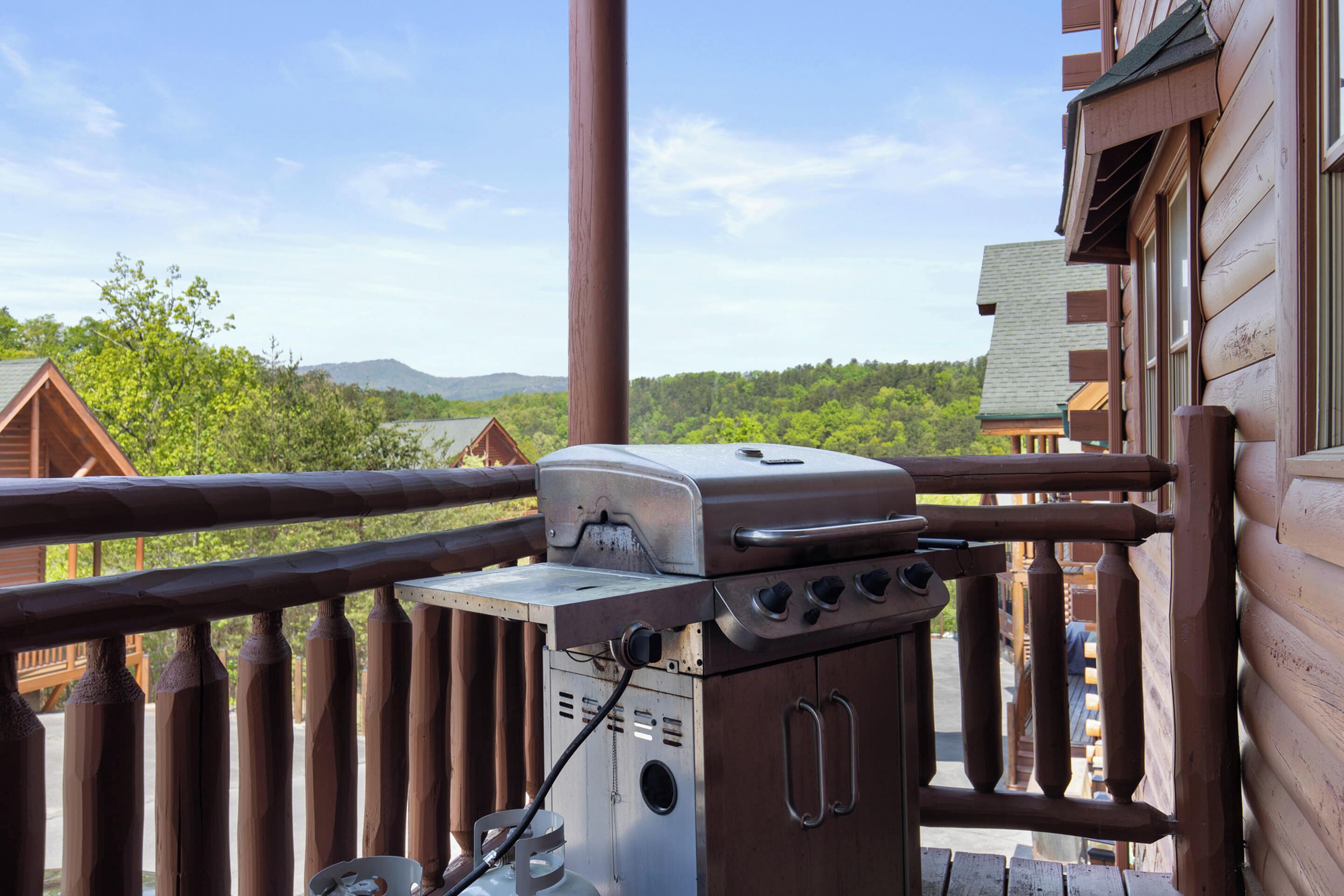 A wooden balcony with a barbecue grill, overlooking a forested area with mountains in the distance.