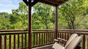 Um deck de madeira com guarda-corpo de madeira, uma única cadeira Adirondack e vista para uma área arborizada.