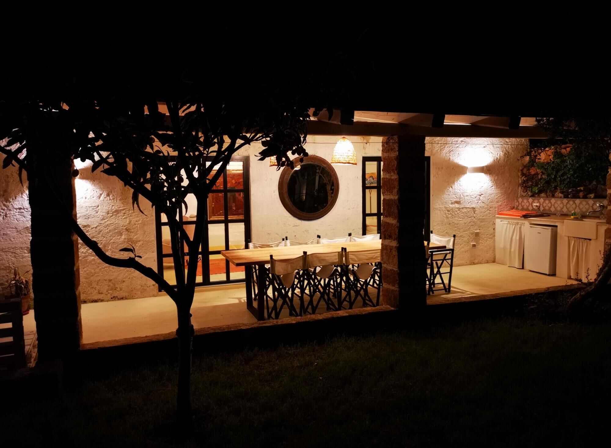 An outdoor dining area with a table and chairs, a round mirror on the wall, and a kitchen area with a sink and cabinets.