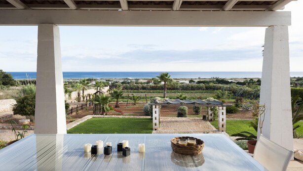A terrace with a view of the sea, a garden, and a table with candles and a basket.
