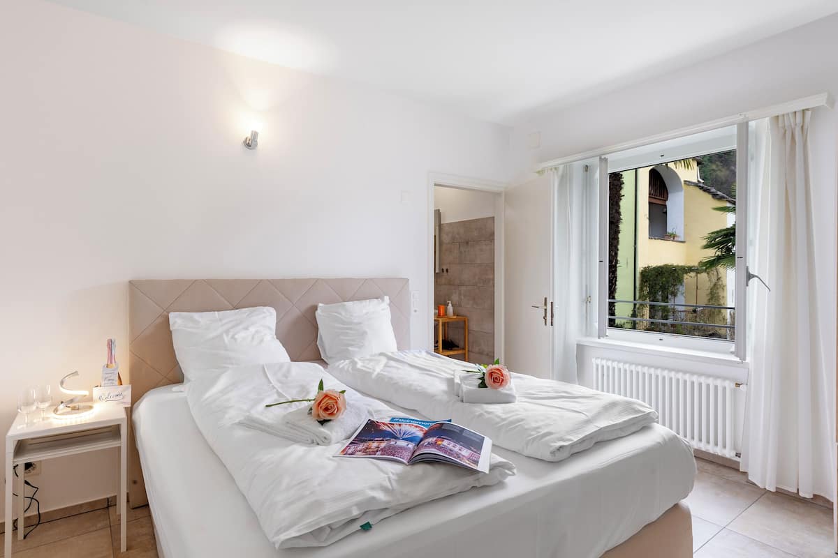 Habitación de hotel con dos camas, una mesita de noche con lámpara, una ventana con cortinas y vistas al exterior.