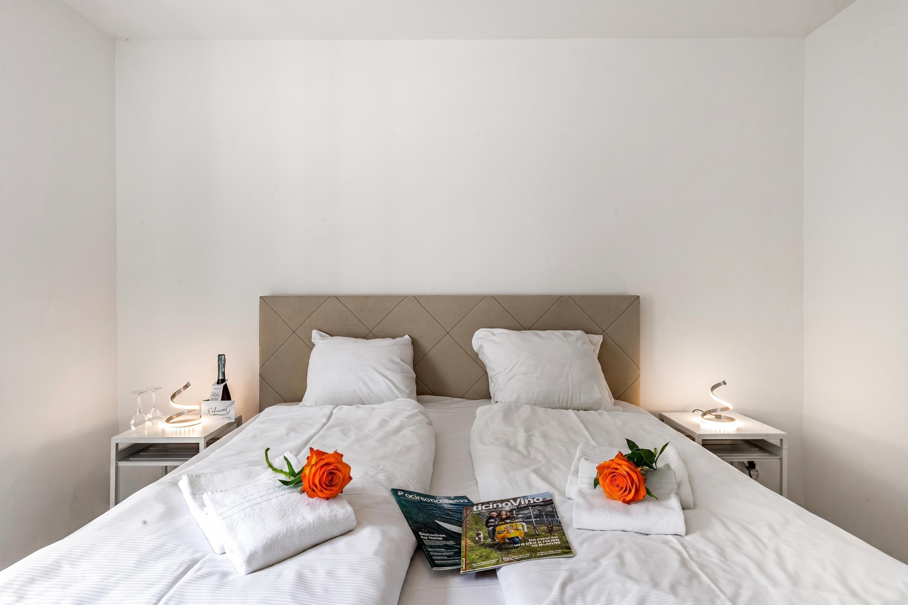 A neatly made bed with white linens, two pillows, and two rose bouquets. There are two bedside tables with lamps and a magazine on the bed.