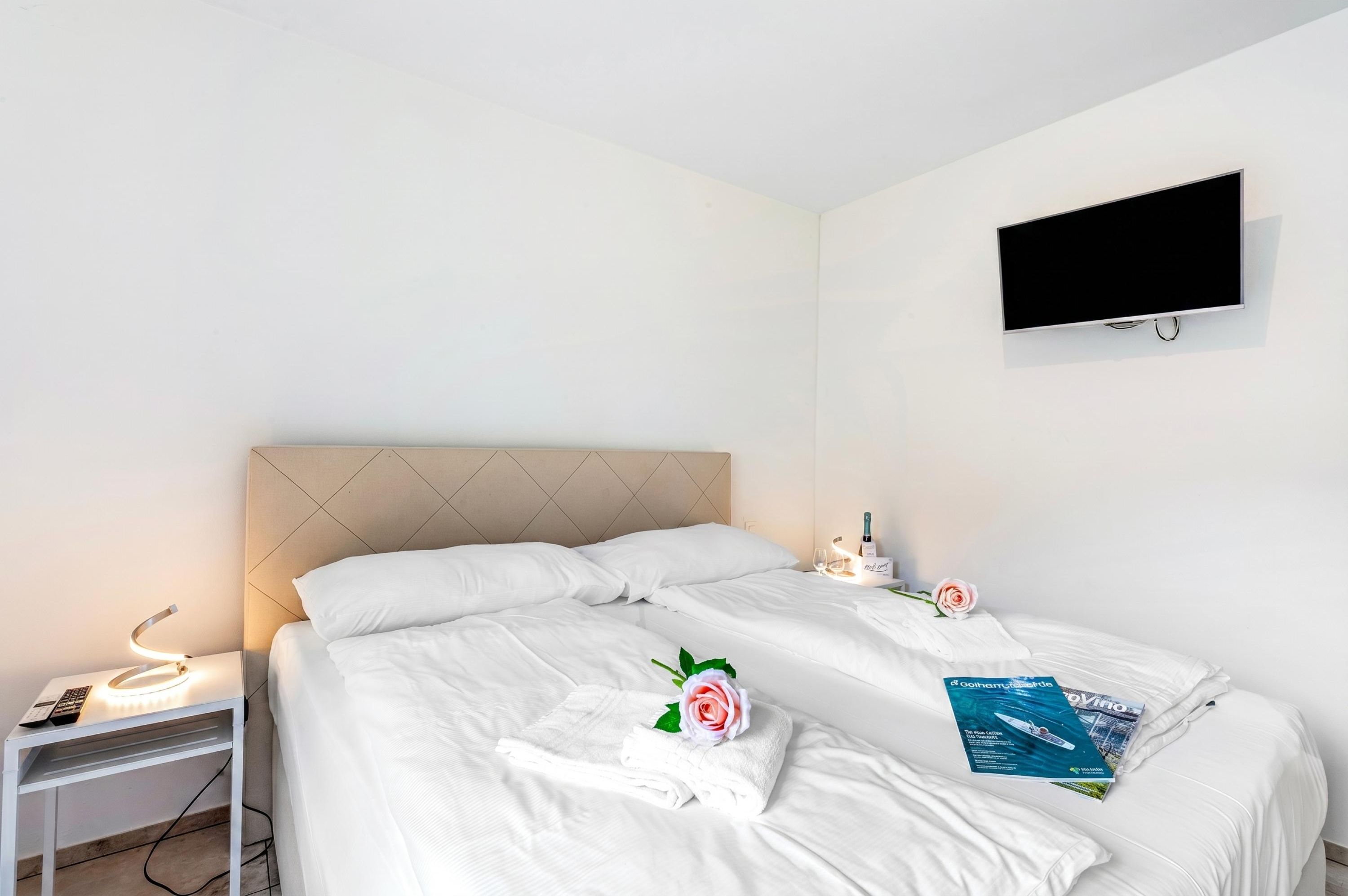 Une chambre d’hôtel avec deux lits, un téléviseur fixé au mur, une table de chevet avec une lampe et un magazine posé sur le lit.