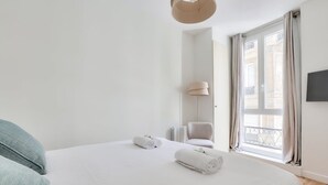 Une chambre avec un grand lit, deux serviettes enroulées, une chaise, une lampe et une fenêtre avec des rideaux.