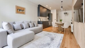 Un salon moderne avec un canapé d’angle gris, un coin repas avec une table et des chaises blanches, et une cuisine avec des meubles de couleur sombre.