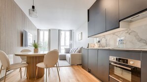 Une cuisine moderne dotée d’un four encastré, un coin repas avec une table ronde et des chaises, et une grande fenêtre laissant entrer la lumière naturelle.