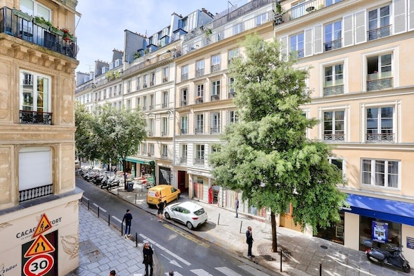 A multi-story building with a row of balconies, a street with parked cars, and a pedestrian crossing.