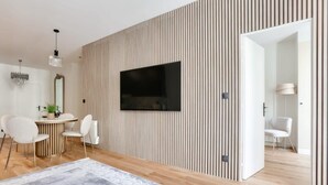 Un salon moderne doté d’un grand téléviseur à écran plat fixé sur un mur en bois à lattes, un coin repas avec une table ronde et des chaises, et un couloir menant à une autre pièce.