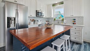 A modern kitchen with white cabinetry, stainless steel appliances, a wooden island, and a view of a blue house through the window. - Birds of Paradise by Ryson Vacation Rentals (Freeport)