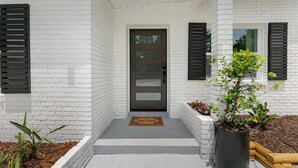 Une maison moderne avec une façade en briques blanches, une porte d’entrée marron foncé et des volets noirs. On peut voir un paillasson à l’entrée et un pot de fleurs sur le côté droit.