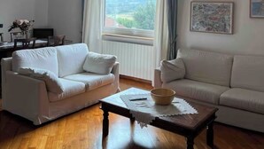 Ein Wohnzimmer mit zwei weißen Sofas, einem Holztisch, einer Pendelleuchte, einem Fenster mit Vorhängen und gerahmten Kunstwerken an der Wand.